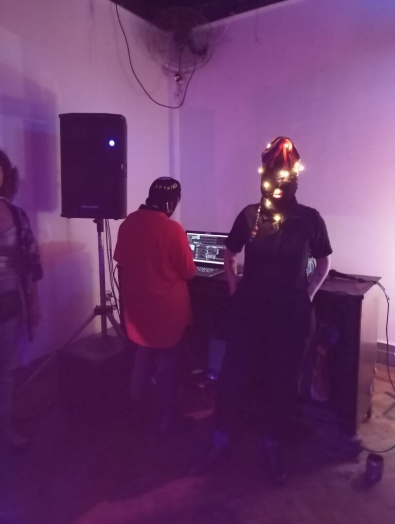 Duas integrantes da Boleia Coletiva junto a um computador e uma caixa de som, sob luz lilás