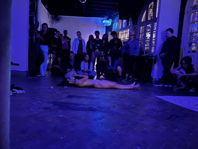 Camila Florentino deitada no chão do salão do Estúdio Lâmina, sob luz azulada, com o público ao fundo