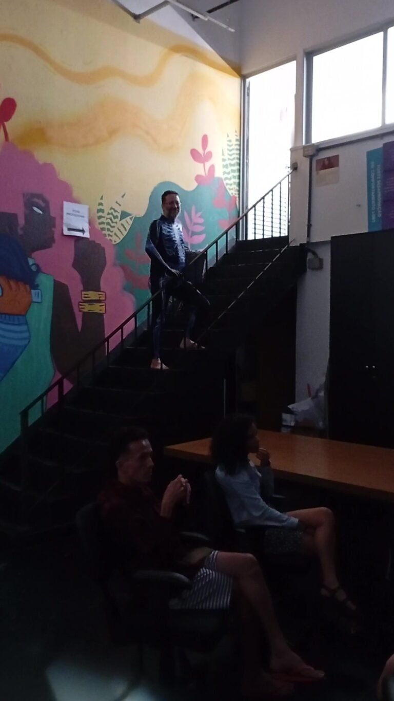 Figura de macacão desce a escada pintada com murais enquanto duas pessoas sentadas acompanham a cena