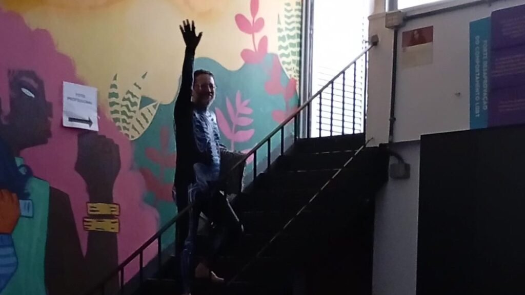 Figura de macacão estampado acena com o braço erguido no meio de uma escada, diante de um mural colorido