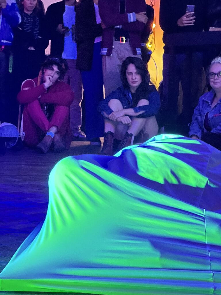 Escultura vestível de malha elástica no chão, em azul e verde sob luz negra, com o público ao fundo