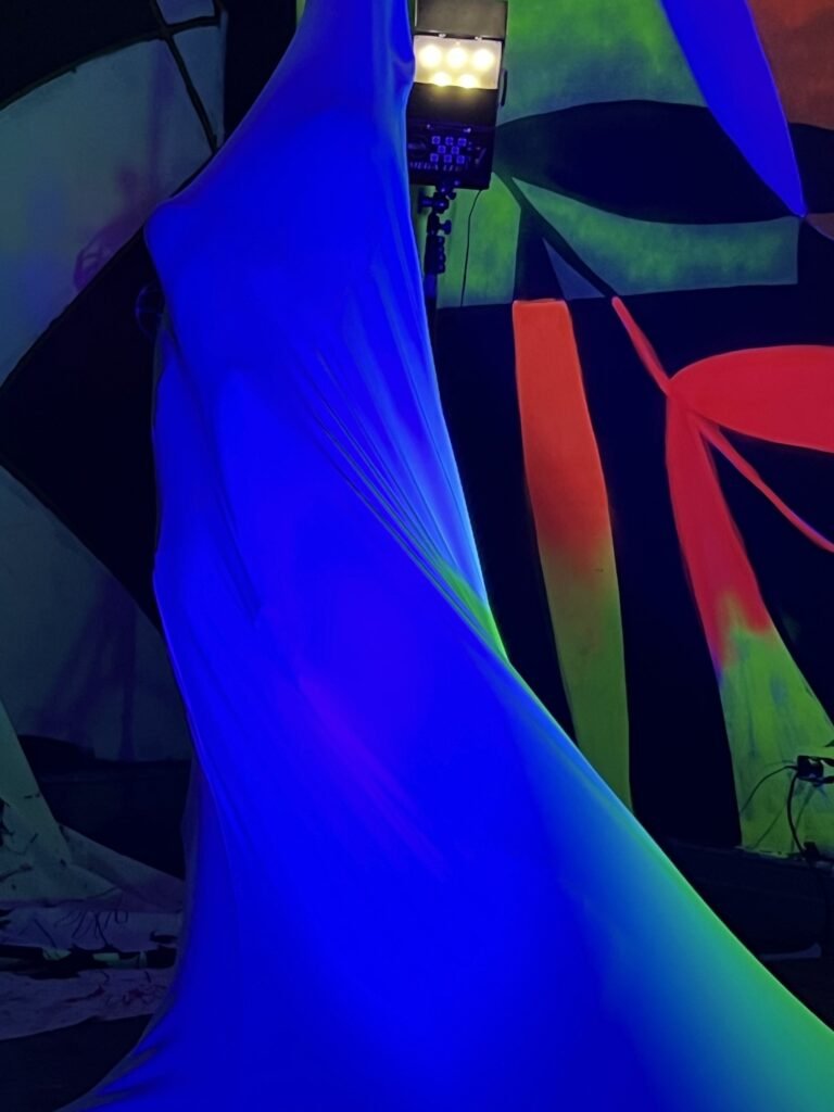 Escultura vestível de malha azul erguida e esticada diante dos murais fluorescentes