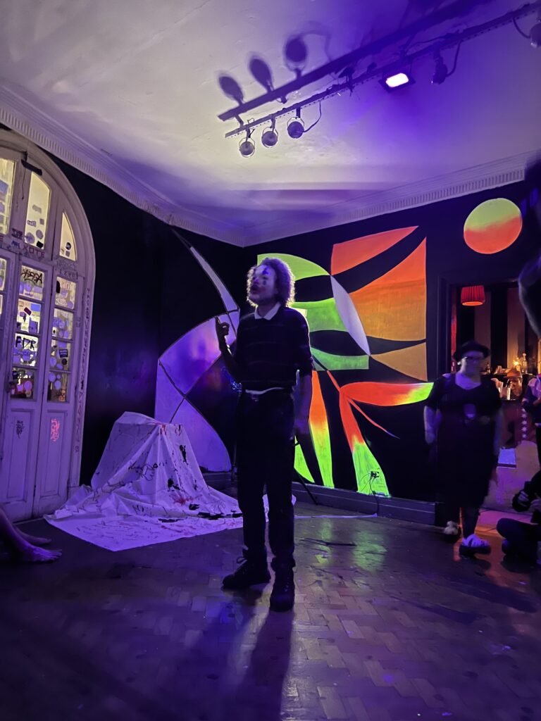 IsTo, com o rosto pintado de palhaço, de pé no salão do Estúdio Lâmina diante dos murais fluorescentes, sob luz roxa