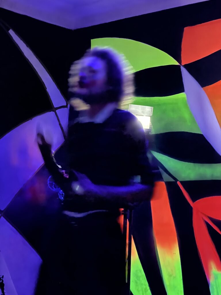 IsTo em movimento, de perto, com as mãos junto ao peito diante do mural fluorescente do Estúdio Lâmina