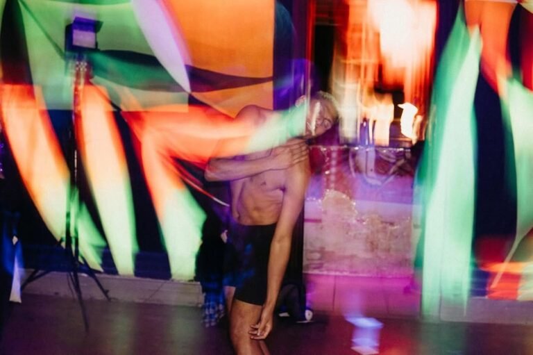 Jon Diegues dança sem camisa diante dos murais fluorescentes, com rastros de luz