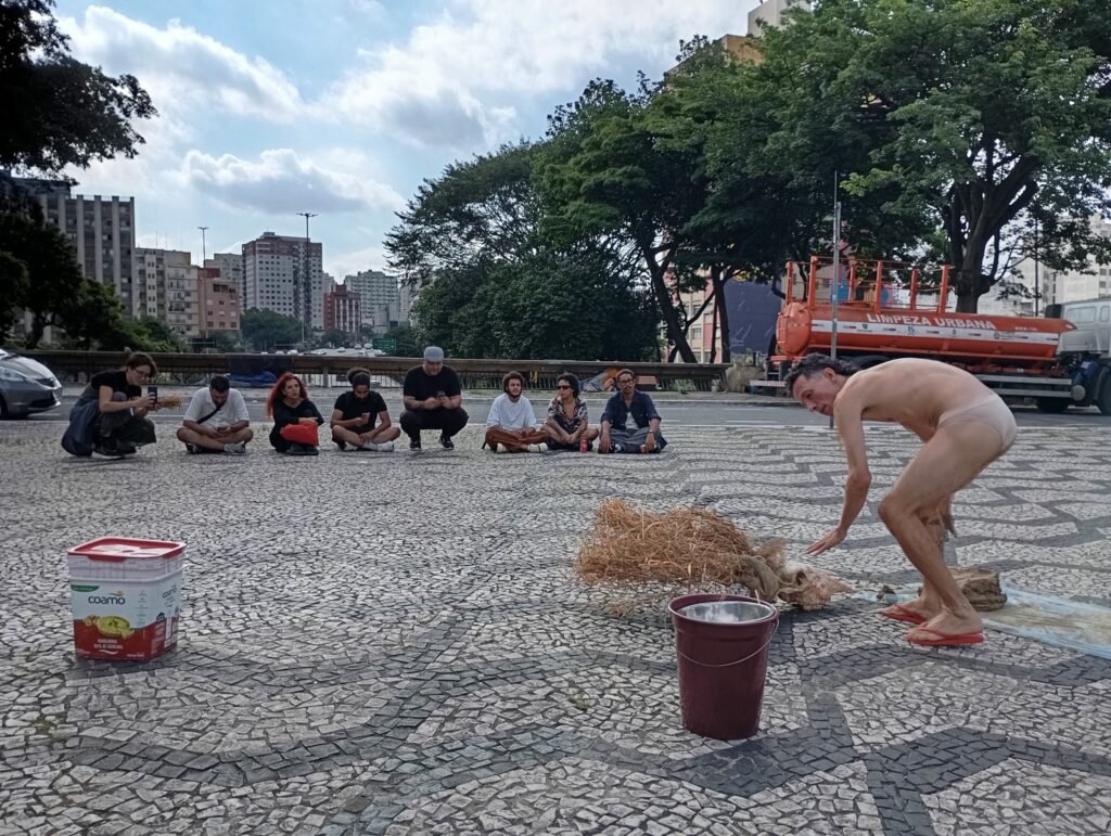 Michel Arantes, de costas e curvado, junto a um feixe de palha e baldes sobre a calçada de pedras portuguesas, com o público sentado ao fundo