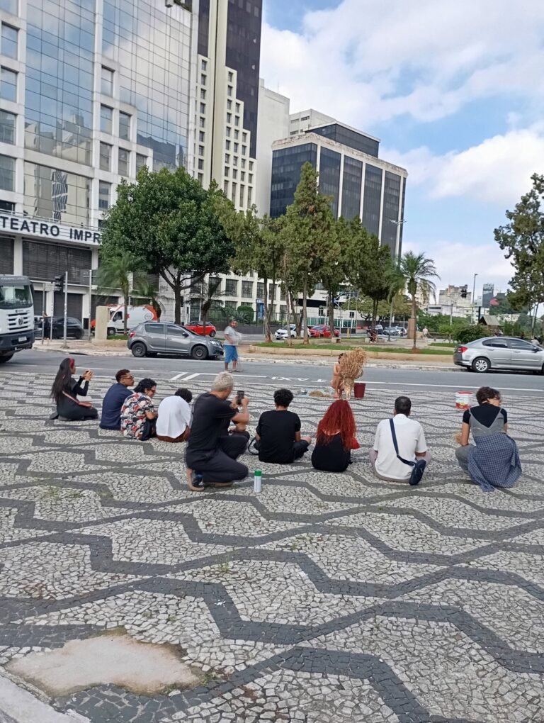 Público sentado na calçada de pedras portuguesas acompanha a performance de Michel Arantes no Bixiga