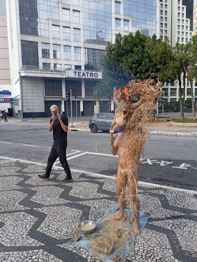 Michel Arantes coberto de palha, fotografado por um homem de preto, diante de um prédio com a placa "Teatro"