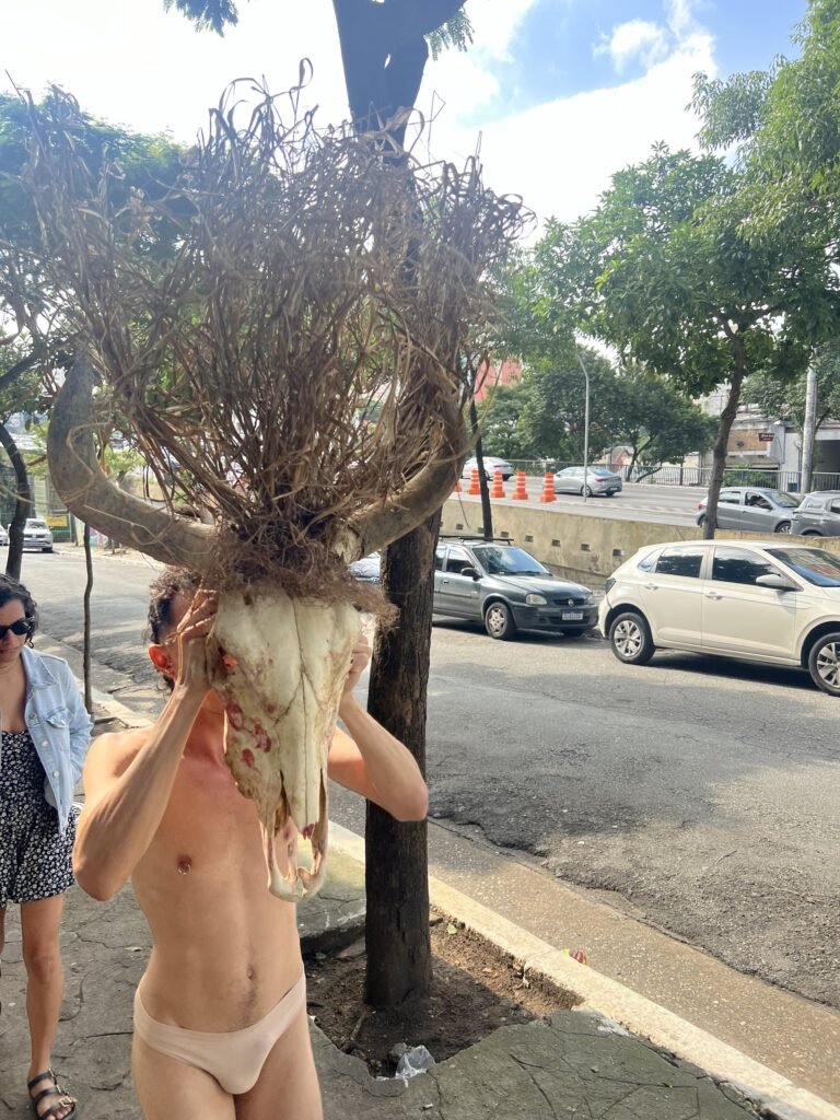Michel Arantes com o rosto coberto por um crânio de animal com chifres e galhos, junto a uma árvore