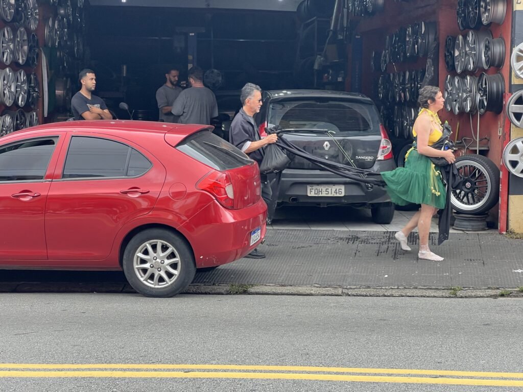 Kareen Labbé Weber, de saia de tule verde e blusa amarela, carrega tecidos pretos diante de uma borracharia em Heliópolis