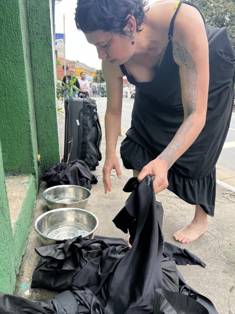 Kareen Labbé Weber, descalça, agachada junto a bacias de metal e tecidos pretos na calçada