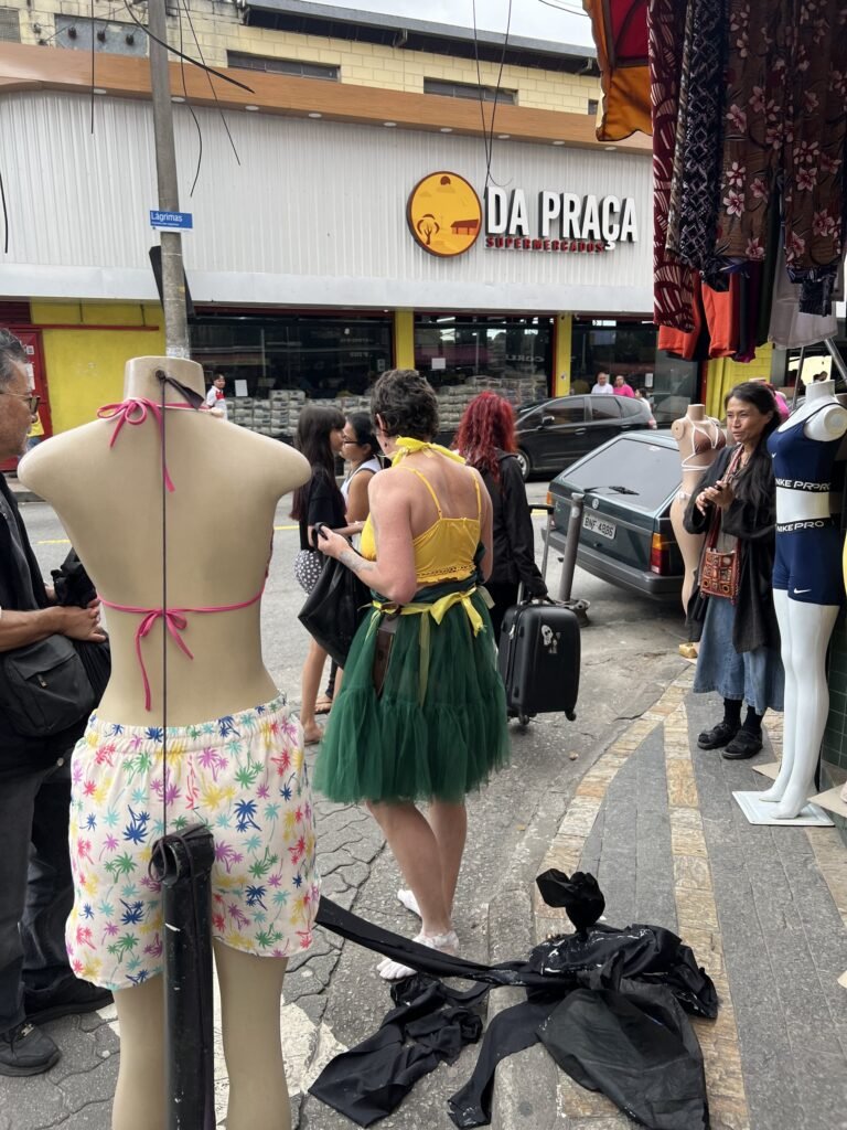 Kareen Labbé Weber entre manequins na calçada de uma loja de roupas, com tecidos pretos no chão