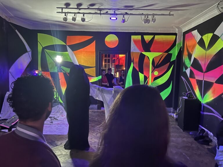 Figuras vestidas de chiffon preto e de chiffon branco se movem pelo salão do Estúdio Lâmina sob luz negra