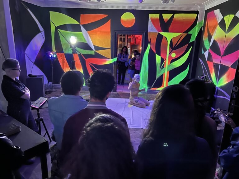 Público em volta do tecido cru onde Reyan Perovano performa, no salão de murais fluorescentes do Estúdio Lâmina