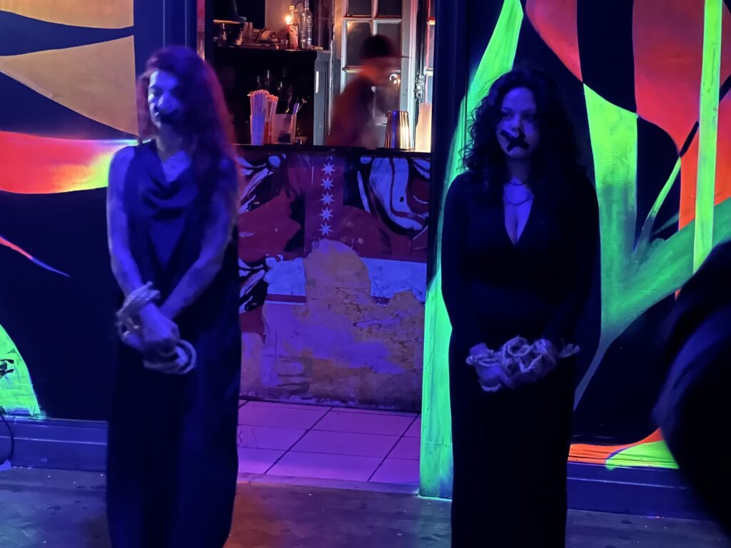 Duas performers de vestido preto, com as mãos atadas, diante dos murais fluorescentes do Estúdio Lâmina