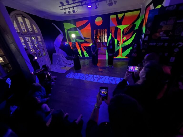 Performers de preto ao fundo do salão do Estúdio Lâmina, com uma faixa de inscrições no chão e o público filmando