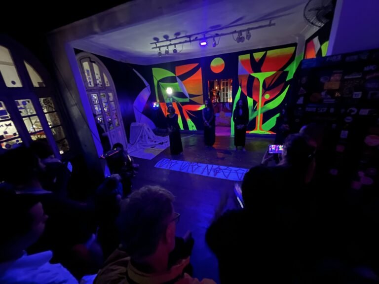 Vista do salão do Estúdio Lâmina com as performers de preto ao fundo e a faixa de inscrições no chão