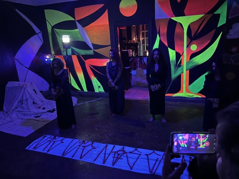 Três performers de vestido preto de pé diante dos murais, com uma faixa de inscrições estendida no chão