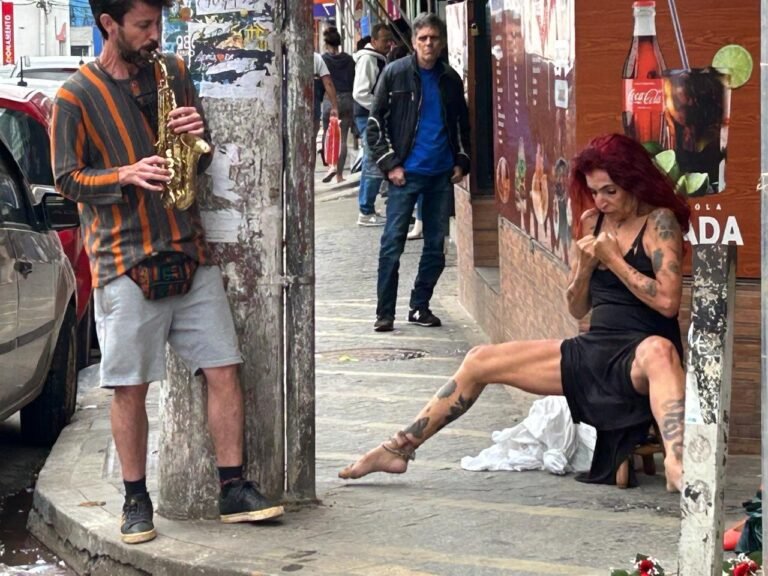 Teresa Ricco sentada num banquinho na calçada, com a perna estendida, enquanto um saxofonista toca ao lado