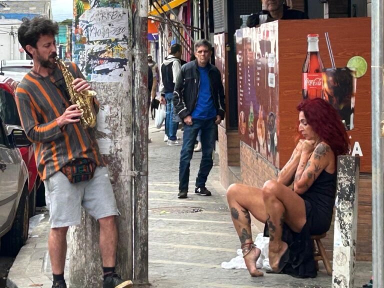 Teresa Ricco curvada sobre o banquinho na calçada, acompanhada por um saxofonista