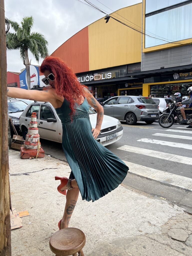 Teresa Ricco apoiada num poste, com um pé sobre o banquinho, diante de uma loja com placa de Heliópolis