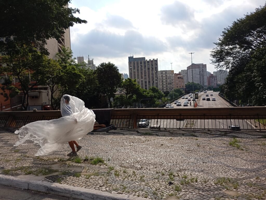 Thales Ferreira envolto numa grande película plástica junto à mureta sobre a avenida