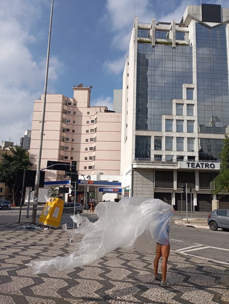 Thales Ferreira arrasta uma longa película plástica pela calçada de pedras portuguesas, diante de um prédio com a placa "Teatro"