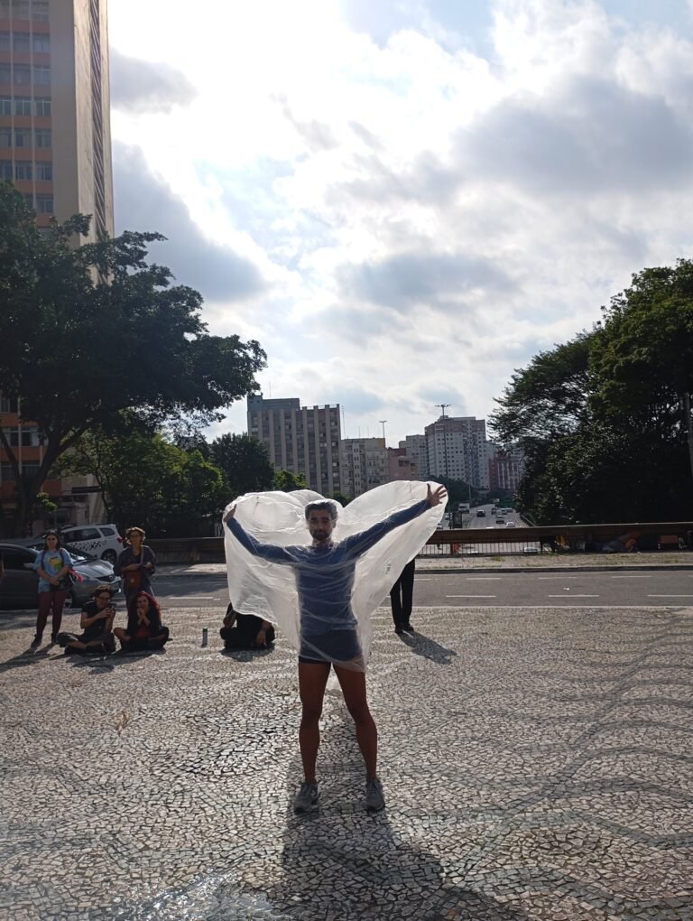 Thales Ferreira abre os braços com a película plástica na praça, com o público sentado ao fundo
