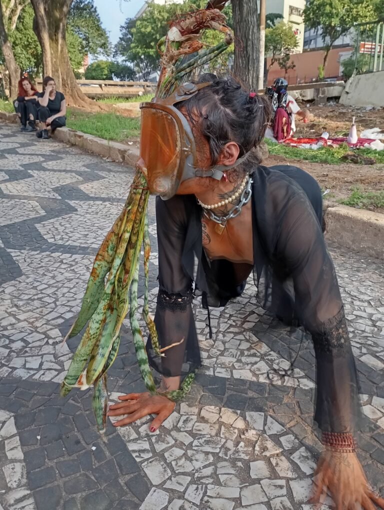Integrante do Triste Midríase de máscara de couro e adereço de folhas, de quatro sobre a calçada