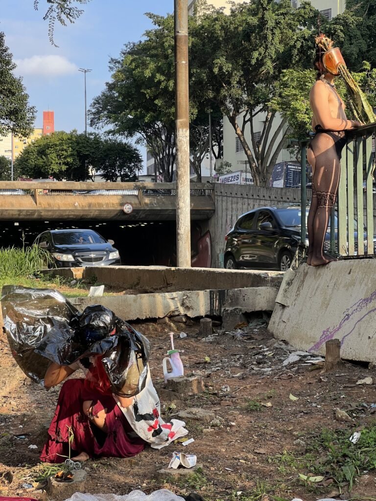 Integrante do Triste Midríase de vermelho junto aos tecidos no chão, com a dupla sobre a mureta ao fundo