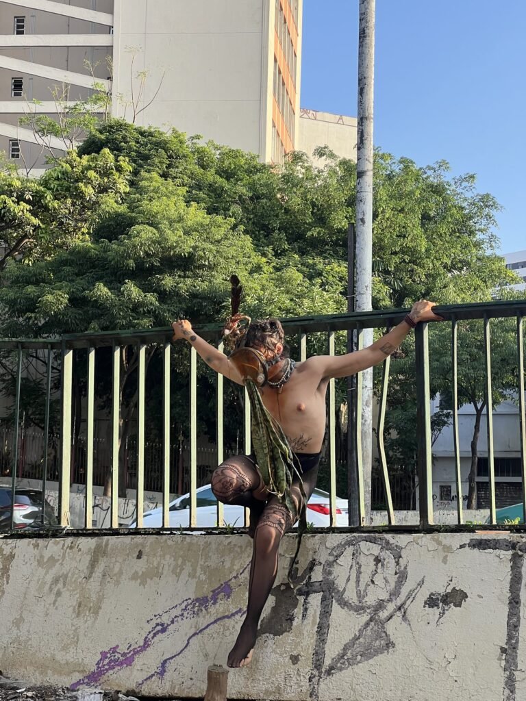 Integrante do Triste Midríase de máscara e adereços pendurado na grade à beira da avenida