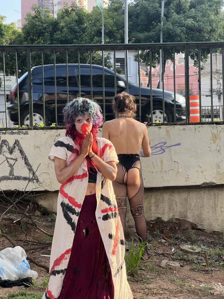 Integrante de nariz de palhaço e casaco branco diante da mureta, com a dupla de costas na grade