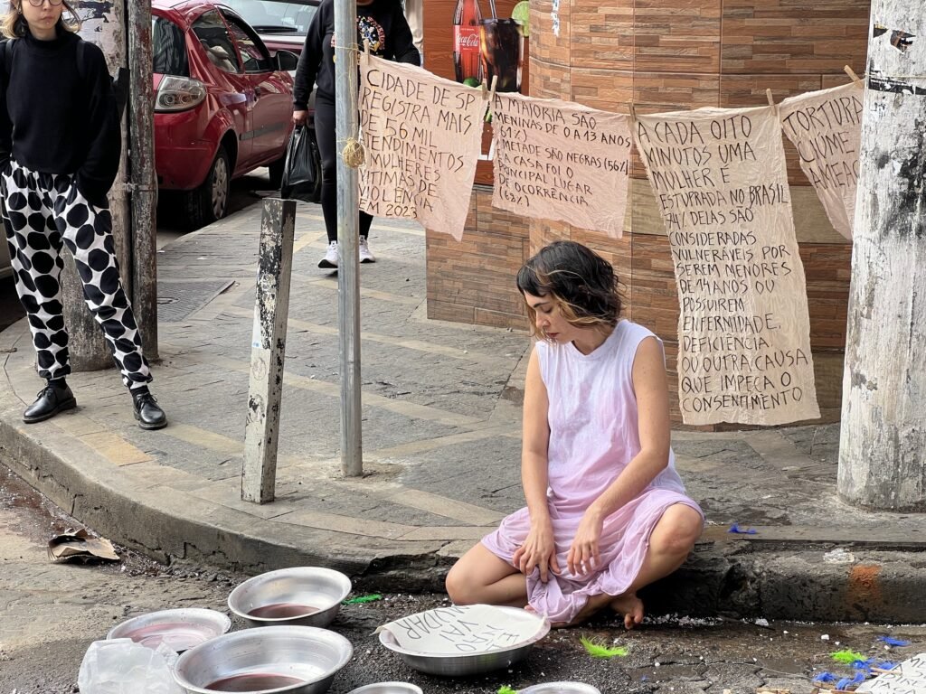 Williana Silva sentada na calçada entre bacias de metal, sob varais de tecidos escritos à mão, numa esquina de Heliópolis