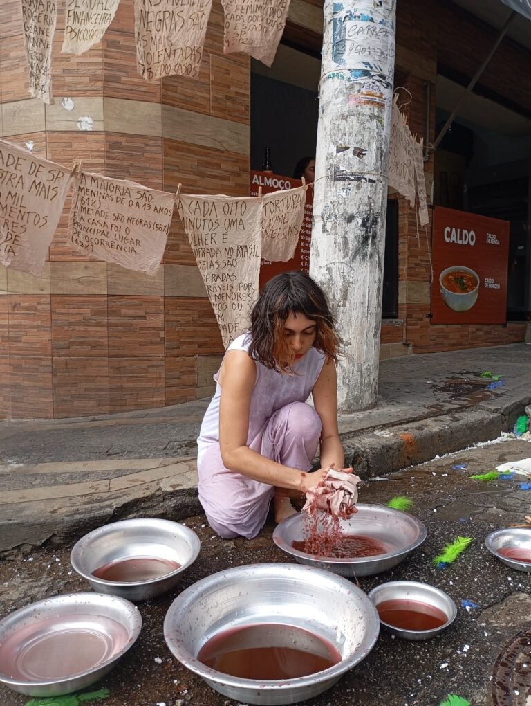 Williana Silva agachada lava as mãos num líquido vermelho entre bacias de metal, diante de tecidos escritos à mão