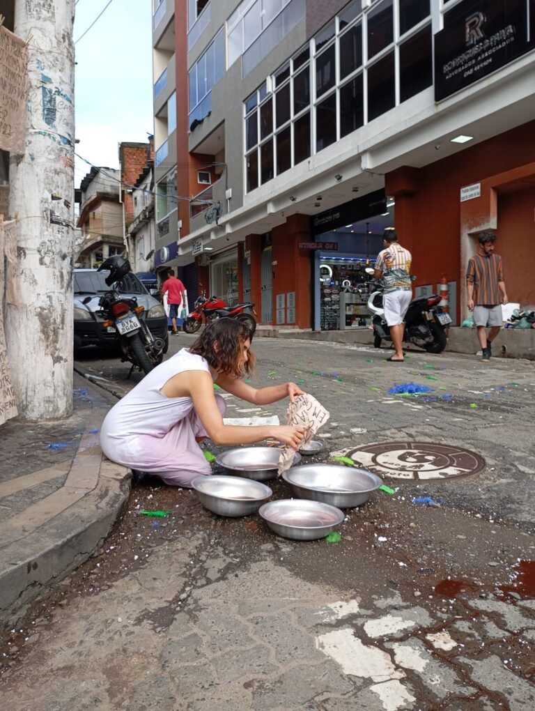 Williana Silva agachada sobre bacias de metal na esquina, com motos e pedestres ao fundo