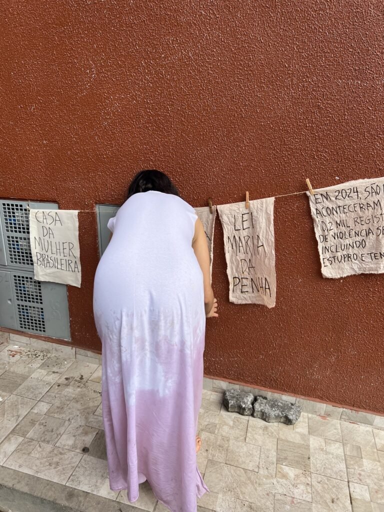 Williana Silva de costas pendura tecidos escritos à mão numa parede vermelha
