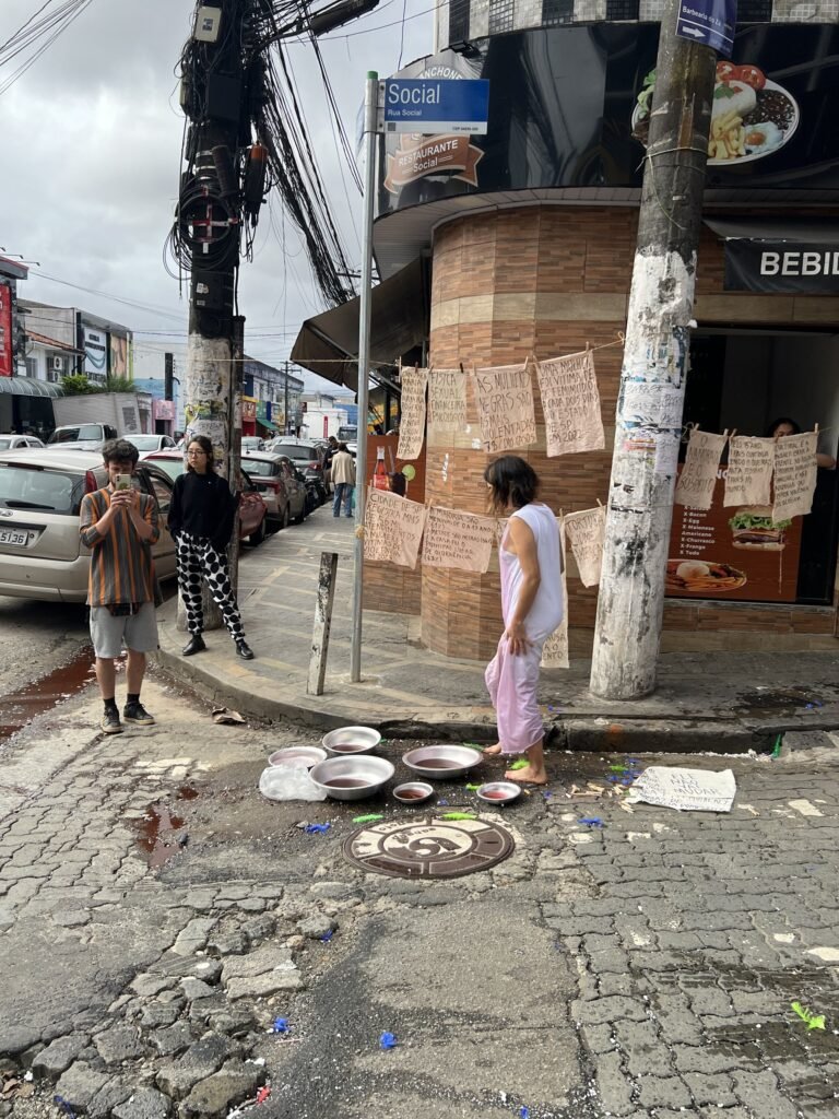 Esquina de Heliópolis com bacias no chão e varais de tecidos escritos, com Williana Silva ao fundo