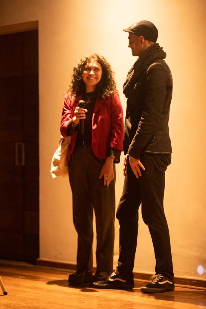Uma mulher com microfone e um homem de boné conversam de pé no palco do auditório da Pinacoteca