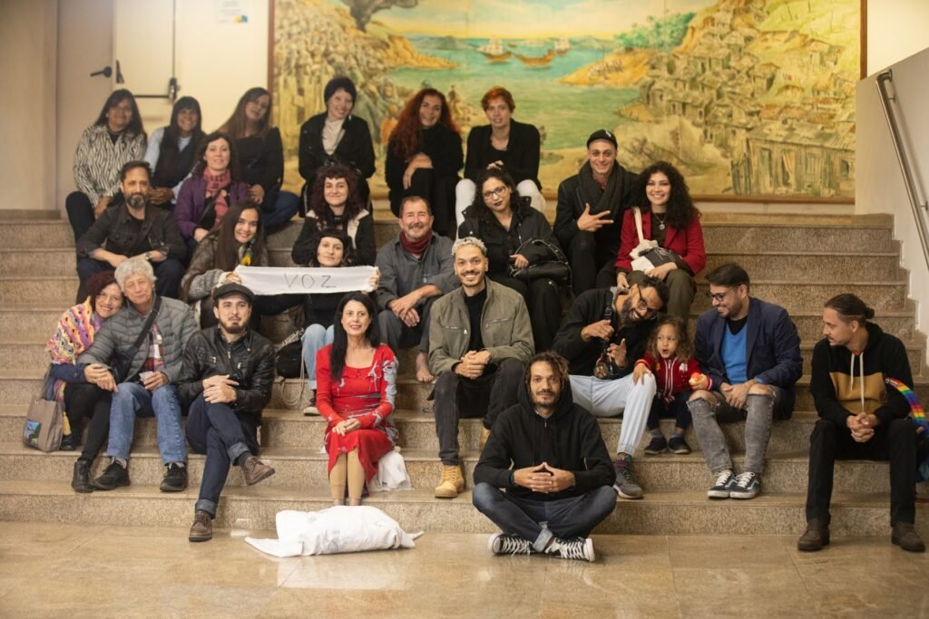 Foto de grupo com artistas, equipe e público sentados na escadaria do saguão da Pinacoteca, sob um grande painel pintado