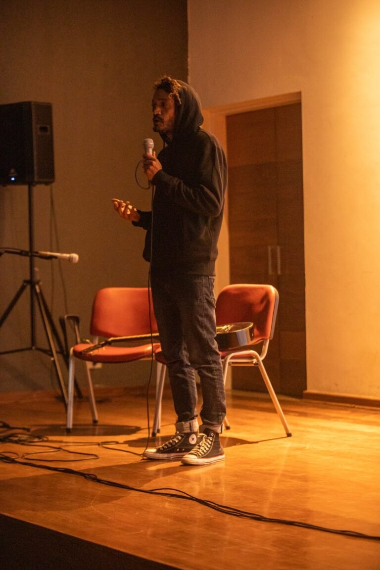 IsTo, de capuz preto, canta ao microfone no palco do auditório da Pinacoteca, sob luz alaranjada