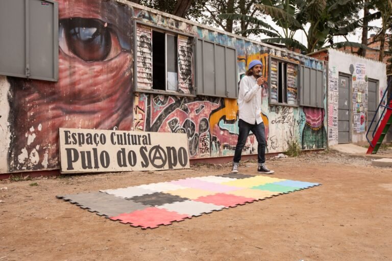 IsTo canta ao microfone diante do muro grafitado com a placa do Espaço Cultural Pulo do Sapo