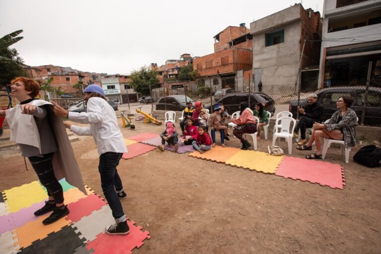 IsTo envolve uma pessoa com o pano branco, sobre os tapetes coloridos, diante do público sentado no pátio do Pulo do Sapo