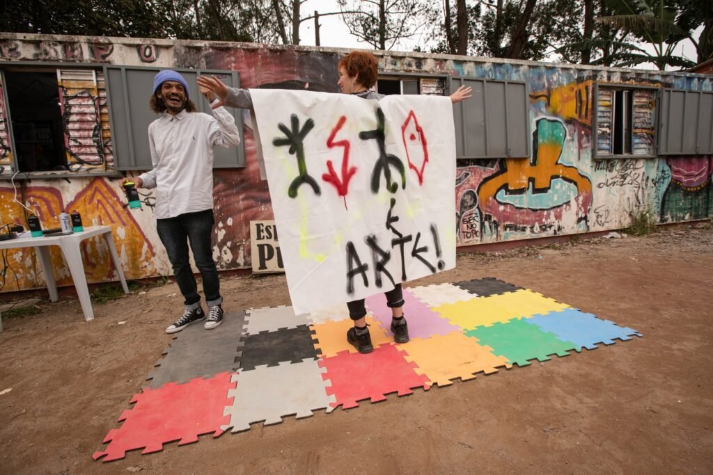 IsTo e outra pessoa seguram o pano branco com os dizeres "IsTo é arte!", sobre tapetes coloridos diante do muro grafitado do Pulo do Sapo
