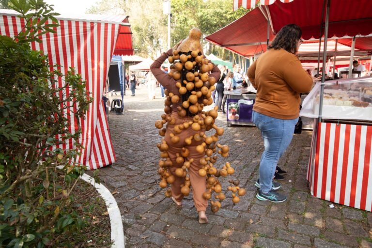 Jéssica Cruz, coberta por cachos de cabaças da cabeça aos pés, caminha entre as barracas listradas da feira do Riacho Grande