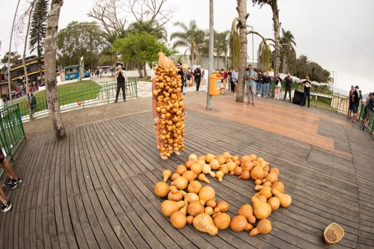Jéssica Cruz, coberta de cabaças, de pé no deck de madeira junto a um círculo de cabaças espalhadas no chão