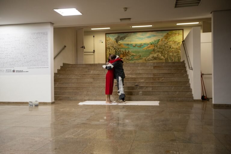 Os dois performers abraçados ao pé da escadaria do saguão, diante de um grande painel pintado e de um pano branco estendido no chão