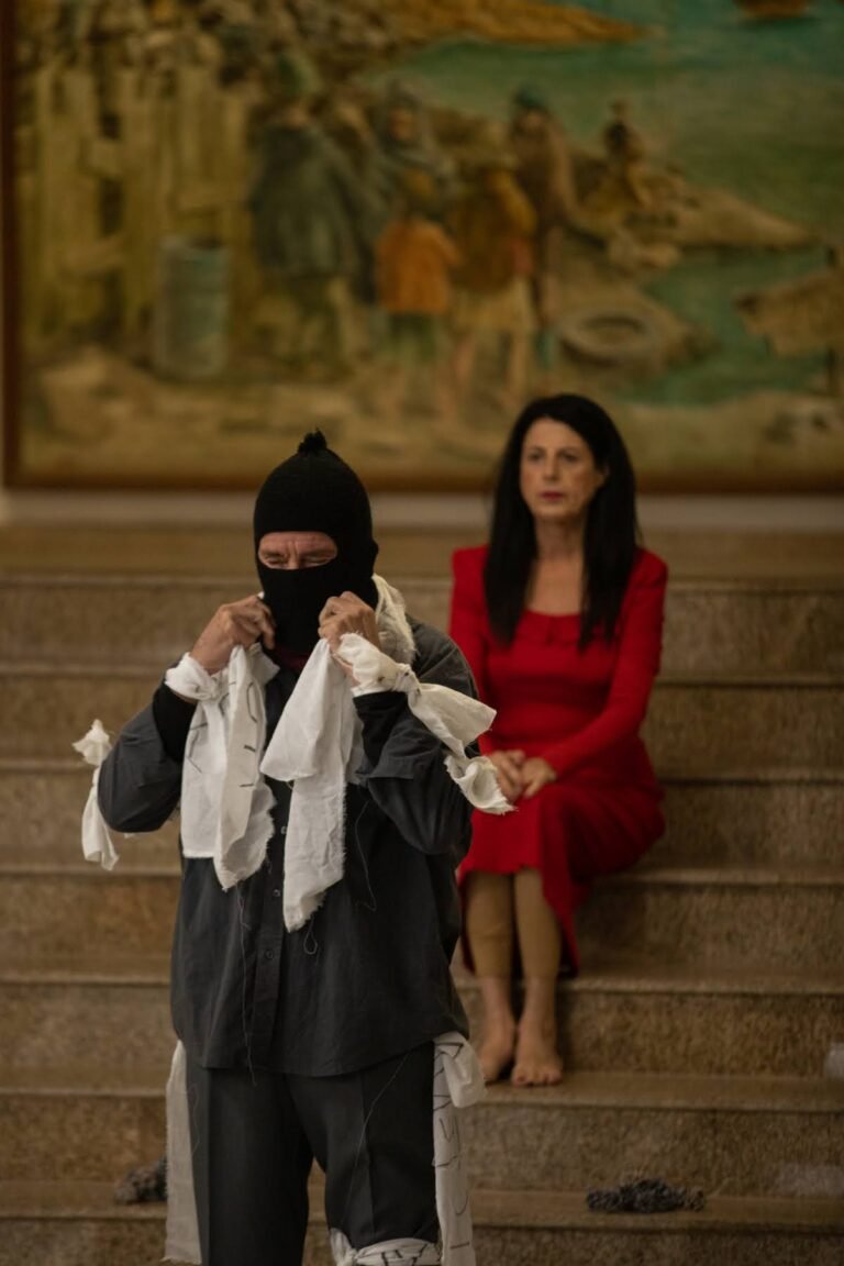O performer encapuzado segura uma tira de pano branco junto ao rosto, ao lado da performer de vestido vermelho sentada na escadaria