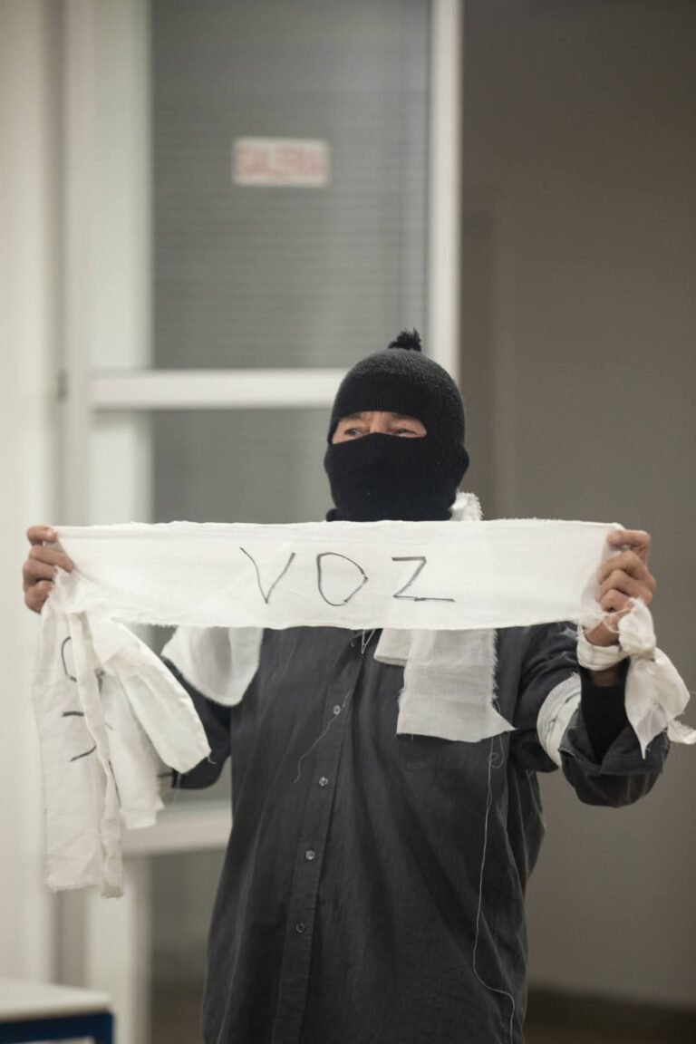 O performer encapuzado ergue uma tira de pano branco com a palavra "voz"
