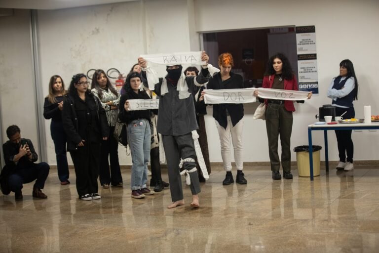 Pessoas do público, de pé, seguram tiras de pano com palavras escritas, ao lado do performer encapuzado