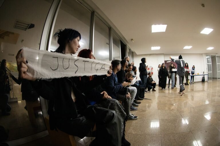 Uma pessoa do público segura uma tira de pano com a palavra "justiça", com a plateia sentada ao longo da parede envidraçada